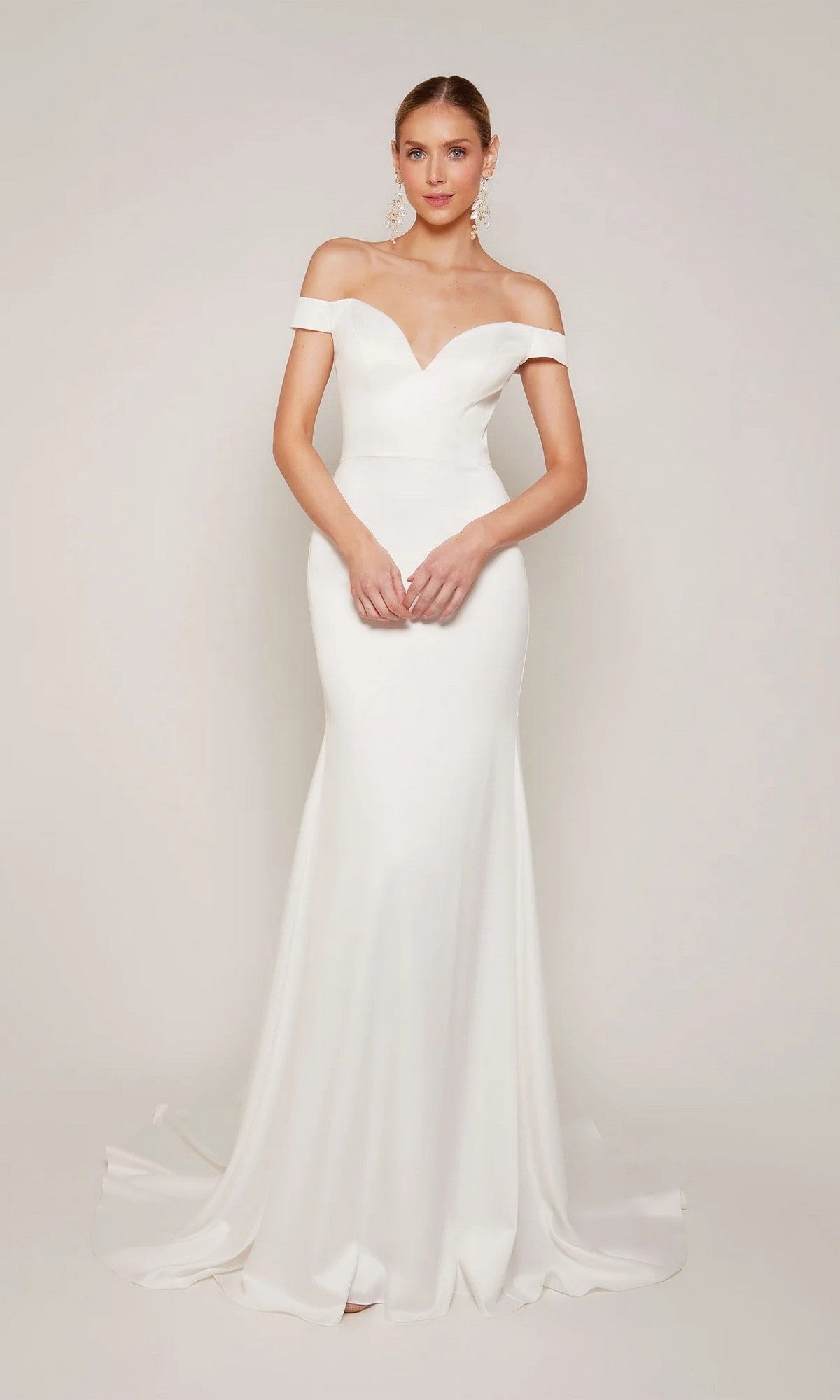 A woman stands before a light background, wearing the Alyce 7098—a white, off-the-shoulder long formal dress featuring a sweetheart neckline, fitted silhouette, and flared floor-length hem.